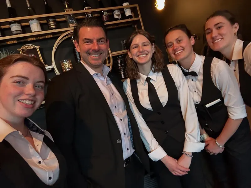 Five Scenic Lounge team members in vests and ties, smiling in front of the wine wall
